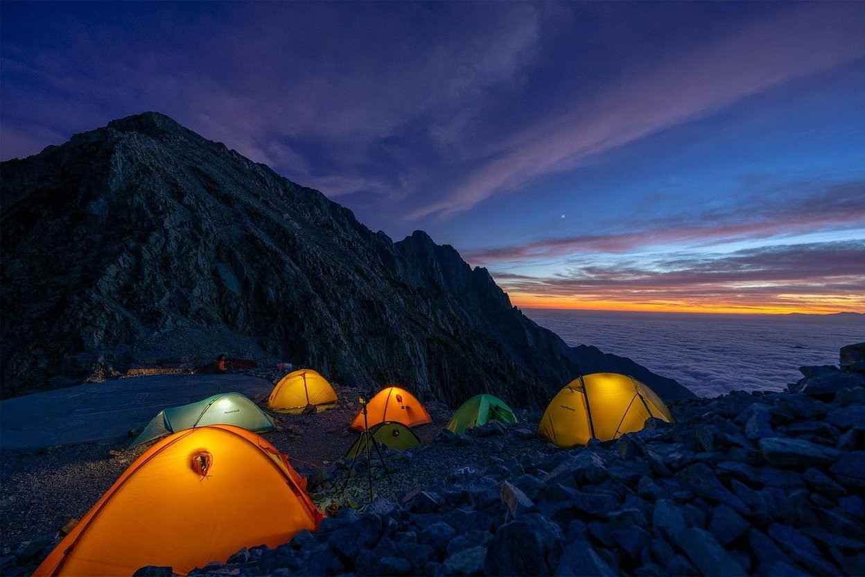 Campsite with glowing tents on a mountain at twilight, clouds below, and a starry sky above.