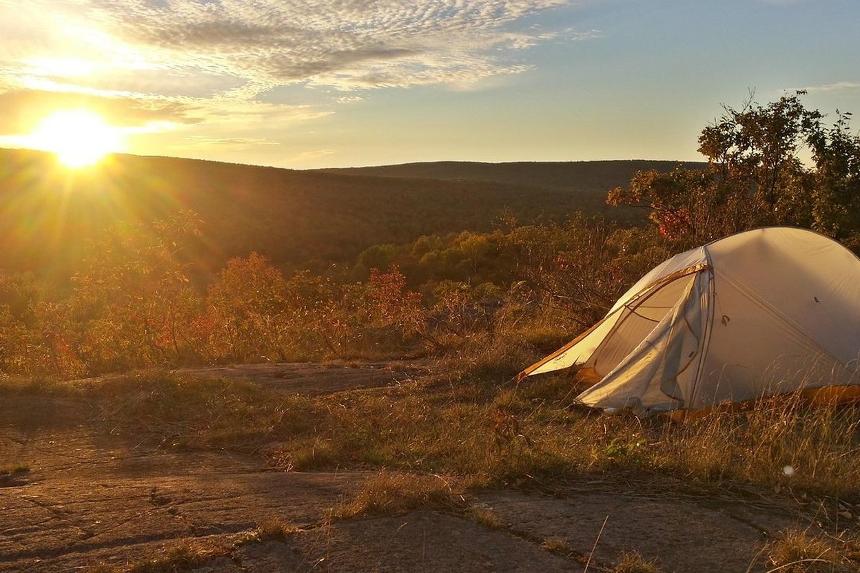 A tent and camp chairs at a fall campsite, with orange-leaved trees and a gravel road in soft late-afternoon light.