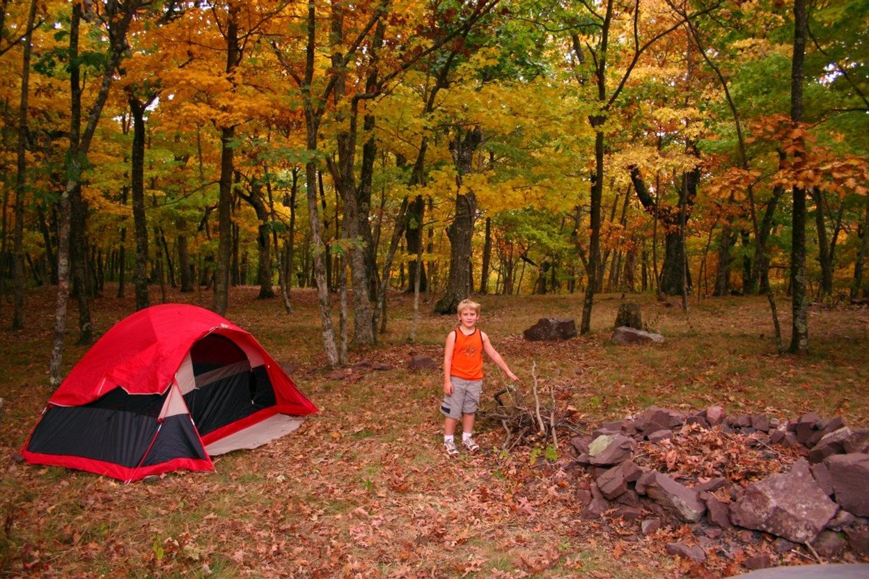 A red tent pitched among orange and yellow fall trees, with a child exploring the campsite