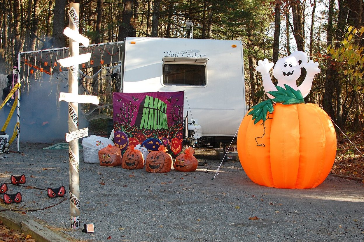 RV campsite decorated for Halloween at dusk, with jack-o'-lanterns on the picnic table, orange string lights on the awning and kids in costumes walking the campground road with candy buckets