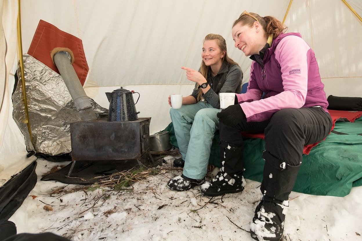 Wood stove with chimney pipe installed inside a canvas hot tent on a cold, snowy evening.