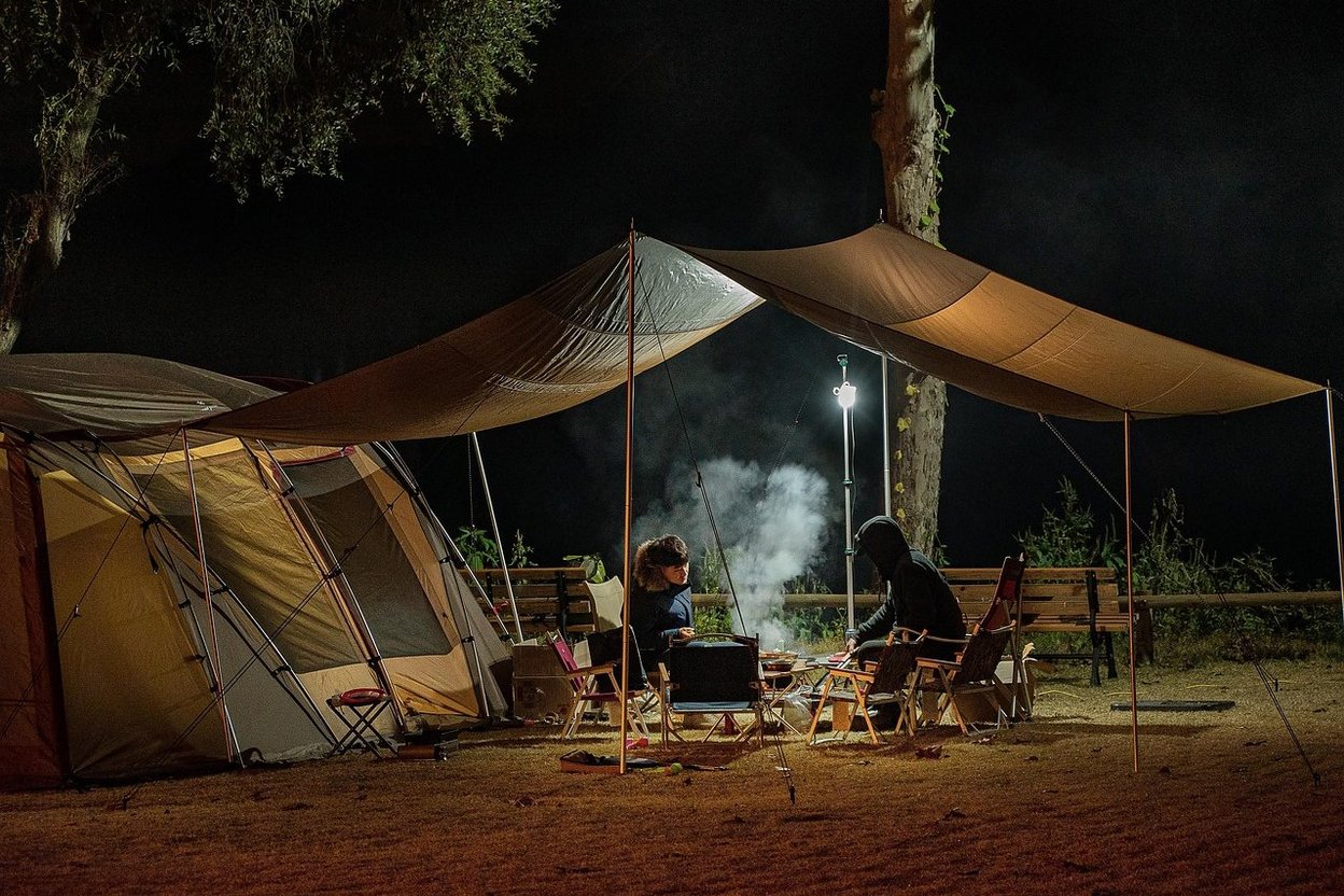 Two people camping at night under a tarp with a tent and campfire.